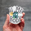 Home body sticker with hand-drawn serif text and a cute little house, with a gray, teal, and yellow color scheme. Hand lettered by Em Dash Paper Co. Printed in the USA on weather-resistant vinyl.