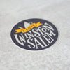 Round vinyl Winston-Salem sticker, hand lettered by Em Dash Paper Co. Grey and yellow, with an illustration of the city's iconic coffee pot.